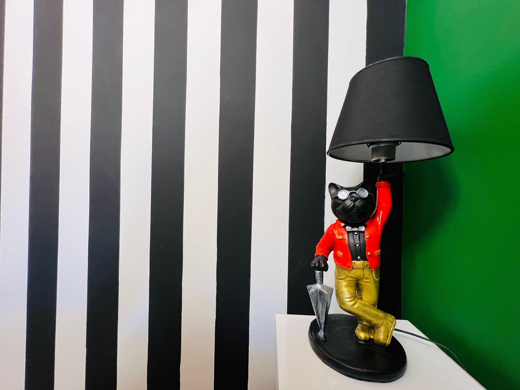 Een zwart-wit gestreepte muur met een zwarte lamp en een beeldje van een kat in een rood shirt en gouden broek.
