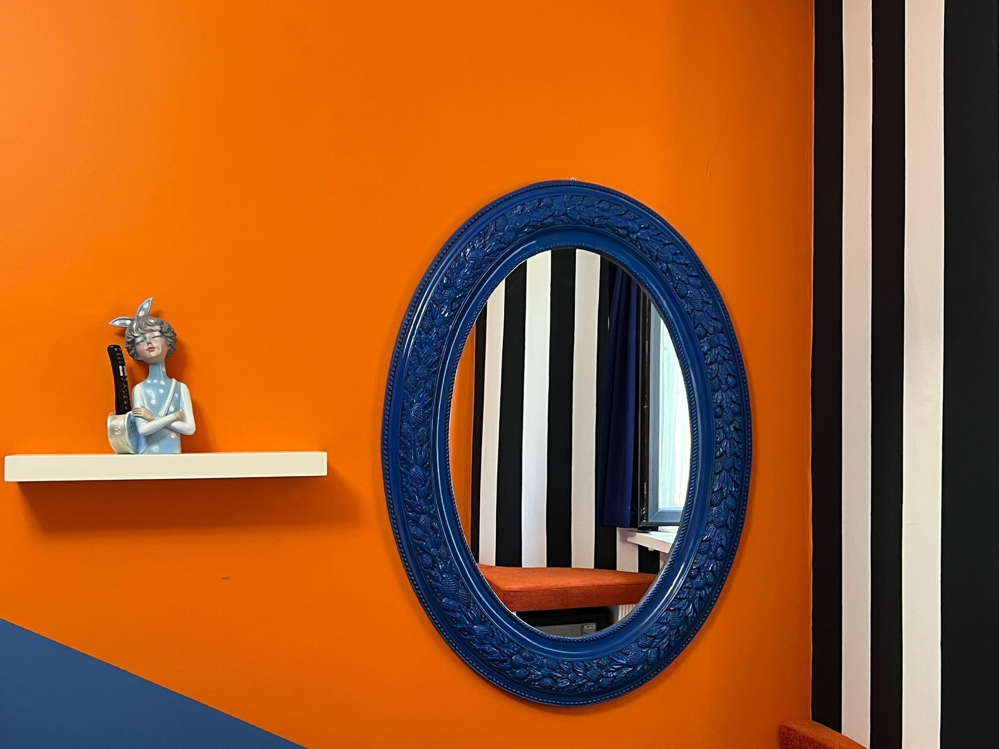 Une pièce avec un mur orange, un miroir à cadre bleu et une étagère blanche sur laquelle est posée une figurine.