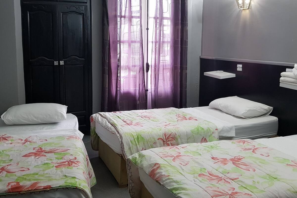 A small, neatly arranged bedroom with two single beds, a dark wooden wardrobe, a window with purple curtains, and a wall-mounted shelf with towels.