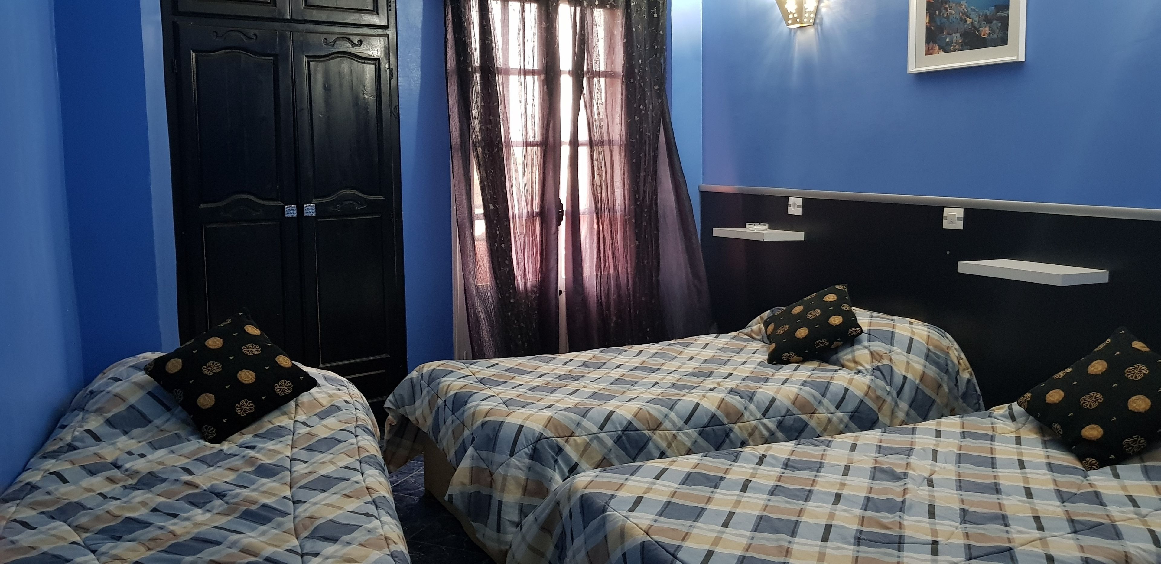 Una habitación con dos camas, un armario negro, una ventana con cortinas translúcidas, una lámpara fijada a la pared y un pequeño estante.