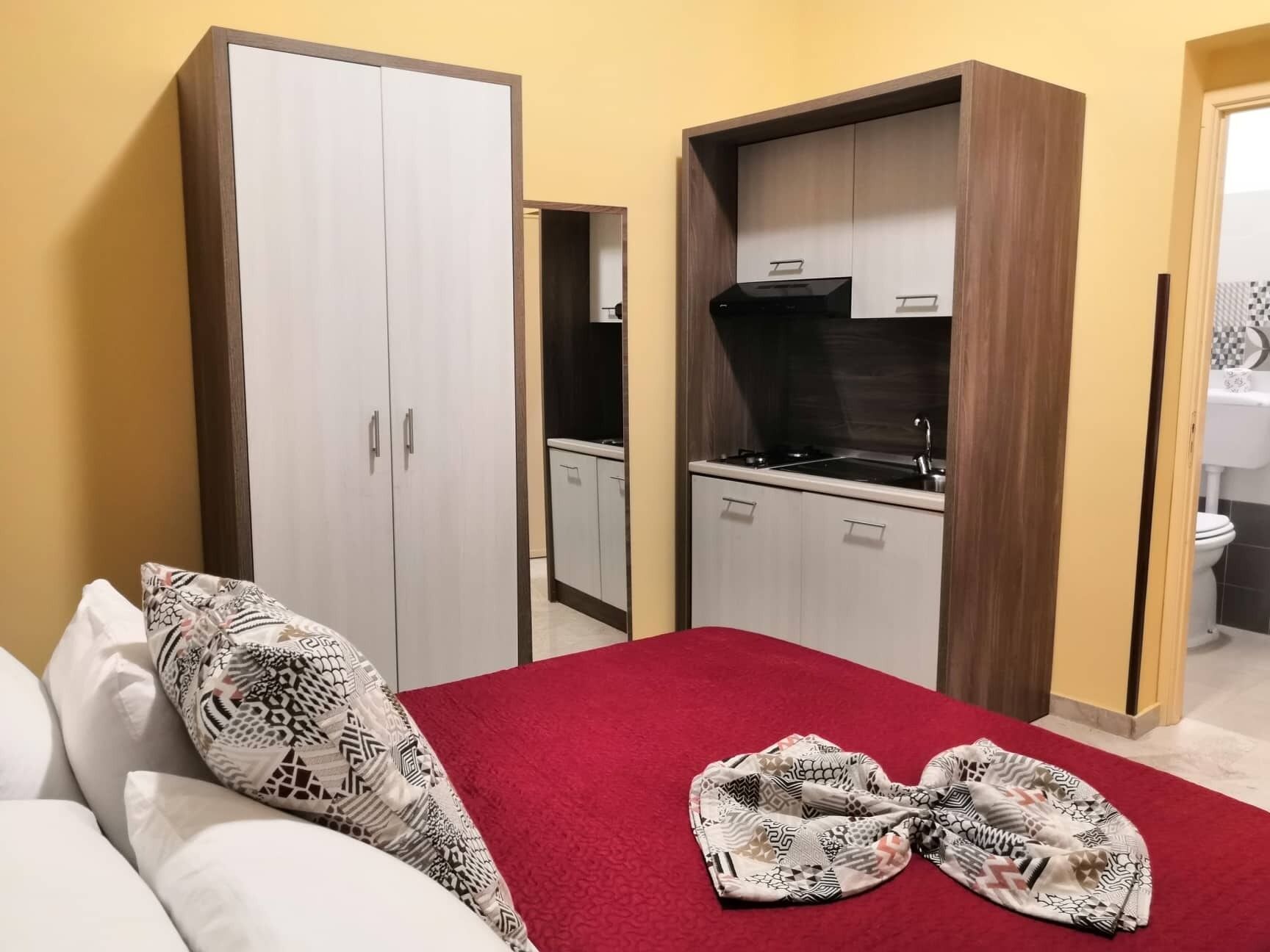 Une chambre à coucher avec un lit, une couverture rouge et un coussin à motifs. On y trouve une armoire avec un miroir, ainsi qu’une petite cuisine équipée d’une cuisinière et de placards.