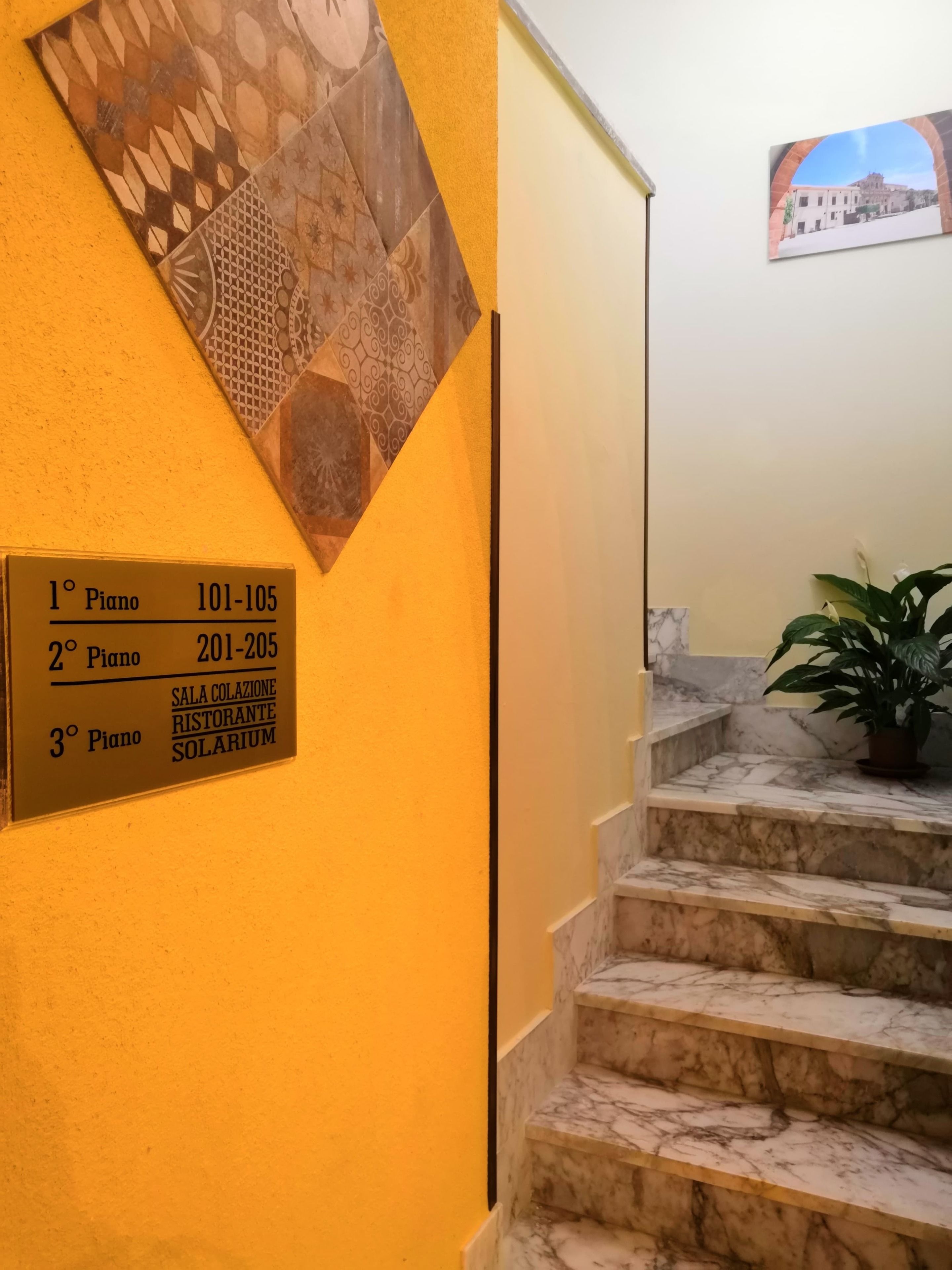 Un escalier avec des marches en marbre menant à un palier. Une pancarte indique les numéros et les noms des pièces : 1er étage, chambres 101 à 105, 2e étage, chambres 201 à 205, 3e étage, salle de petit-déjeuner, restaurant, solarium.