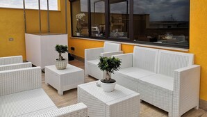 Een terras met witte rieten meubels, potplanten en een groot raam met uitzicht op een woonwijk.