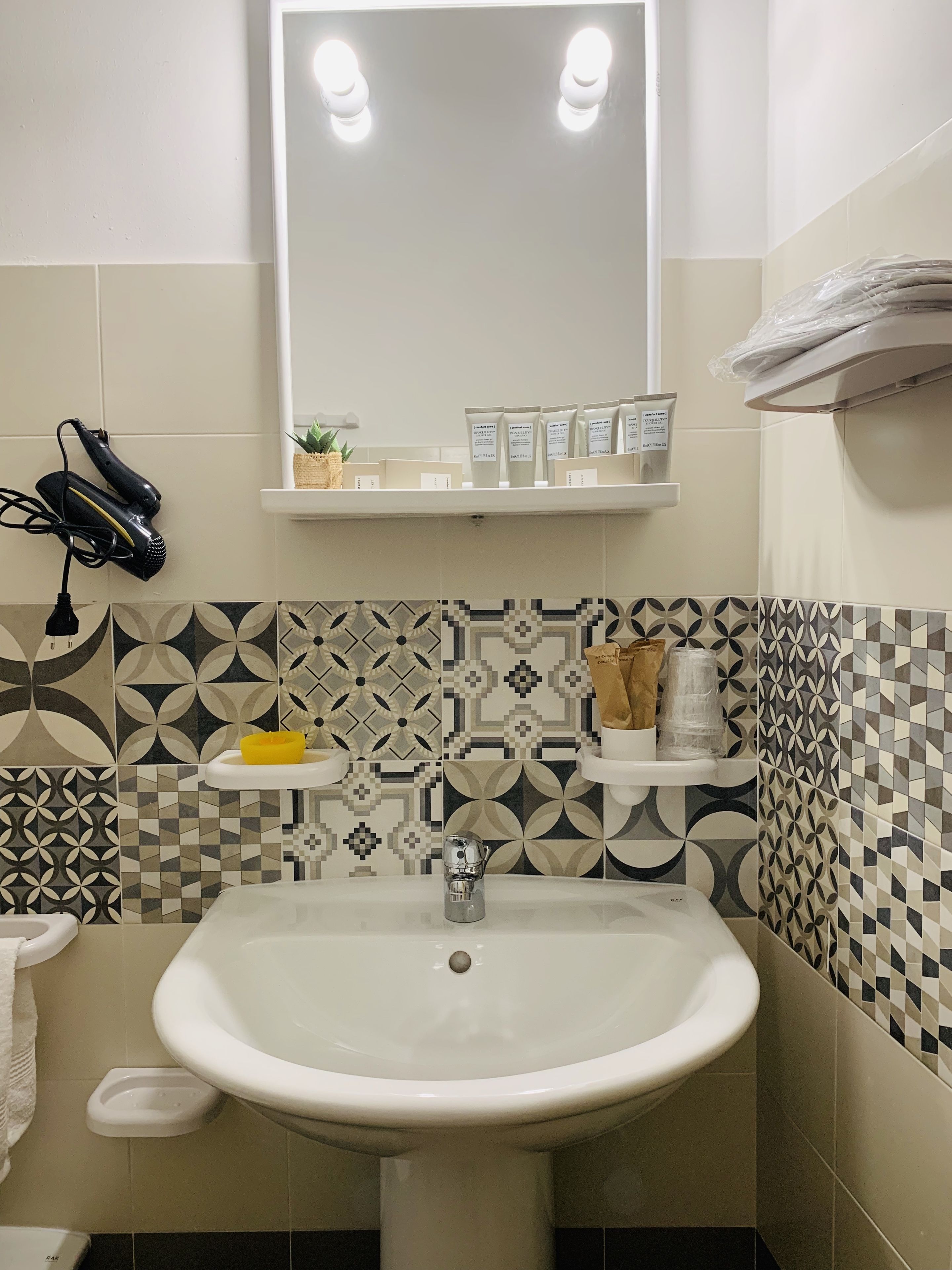 A bathroom with a white sink, a mirror, and patterned tiles. There are toiletries on a shelf and a hairdryer on the wall.