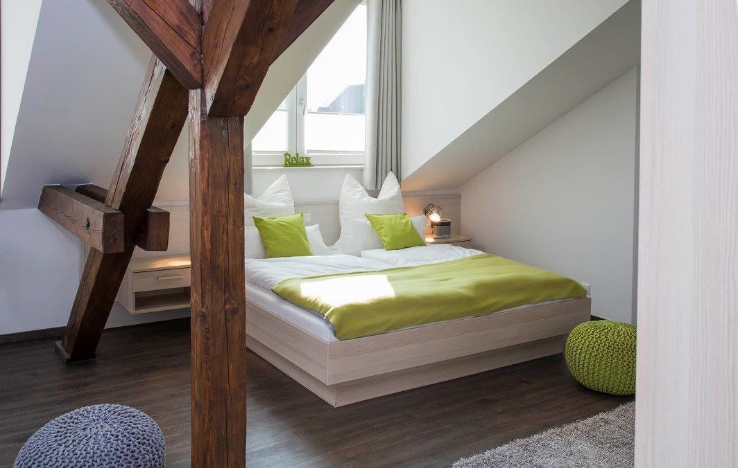 A modern bedroom with a sloped ceiling, a bed with green and white bedding, a bedside table with a lamp, and a window with a view.