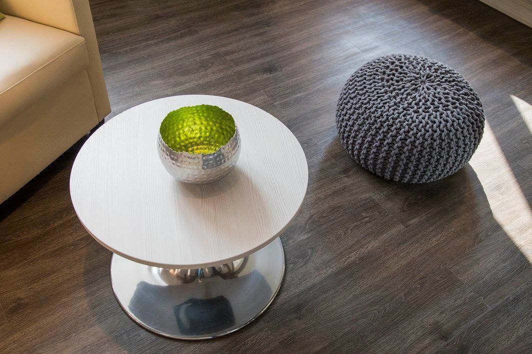 A round white table with a metallic base, a textured green bowl on top, and a grey knitted pouf beside it.