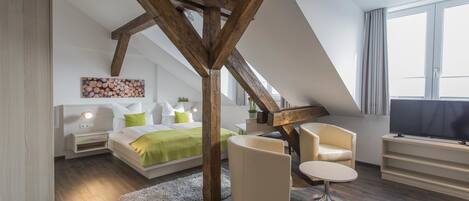 Un dormitorio moderno con techo inclinado, una cama con ropa de cama verde, una zona de estar con una silla y una mesa, y un televisor sobre un mueble.