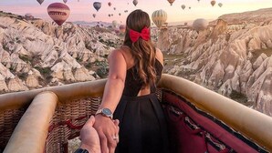 Ein Paar hält Händchen und blickt auf eine Landschaft mit vielen Heißluftballons und markanten weißen Felsformationen.
