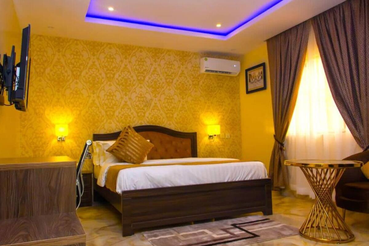 Quarto de hotel com cama de madeira, uma mesinha redonda, televisão fixada na parede e ar condicionado.