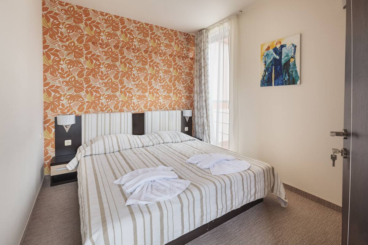 A hotel room with a bed, two bedside tables, a nightstand, a painting on the wall, and a door.