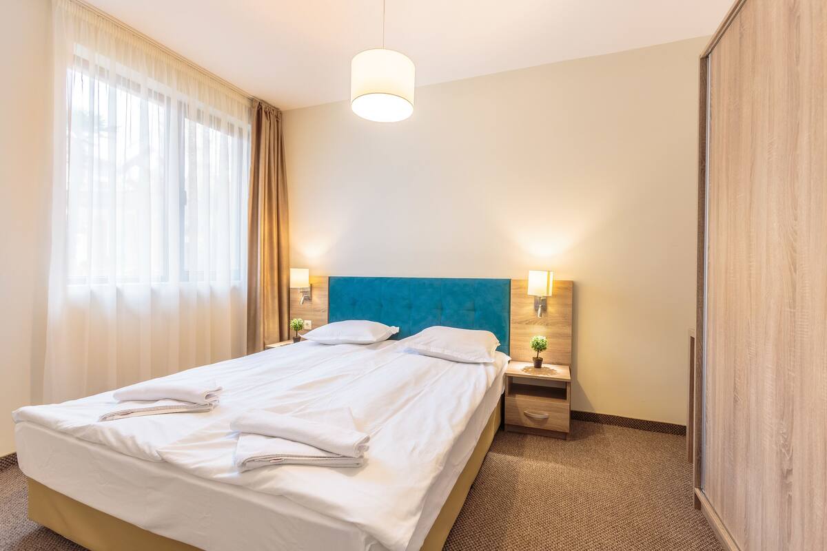 A hotel room with a bed, two bedside tables with lamps, a wardrobe, and a window with curtains.