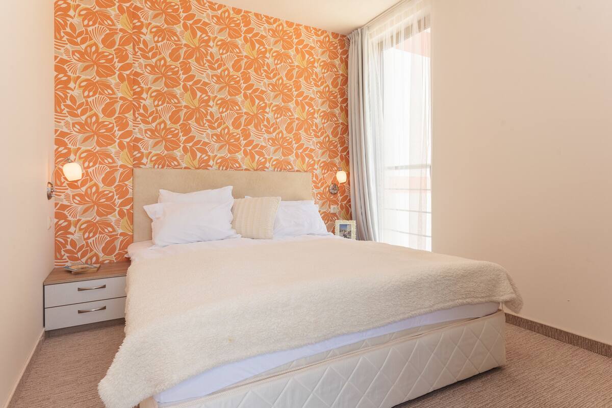 A neatly made bed with a fluffy white blanket, a headboard with a light-colored fabric, and a nightstand with a lamp and a small table.