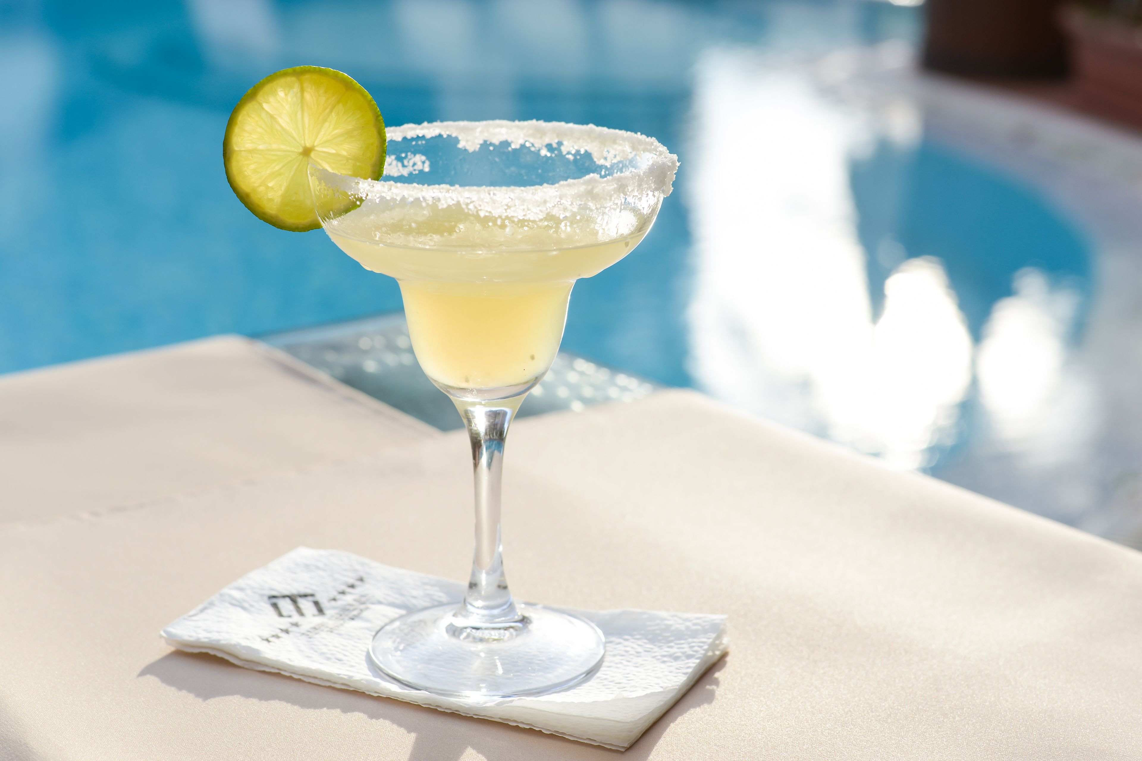 A margarita cocktail with a lime garnish on a cocktail glass, placed on a white napkin beside a pool.