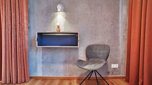 Una habitación con suelo de madera, una silla tapizada de gris, un estante azul con un jarrón y cortinas rojas.