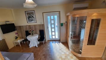 Hotelkamer met houten vloer, een bank, een kleine tafel met een vaas, een aan de muur bevestigde televisie en een deur naar een balkon.