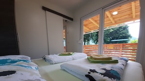 A room with two beds, each with a patterned bedspread and pillows, a wooden ceiling, and a large window with a view of greenery outside. -  Apartment Zonir with sauna (Kobarid)