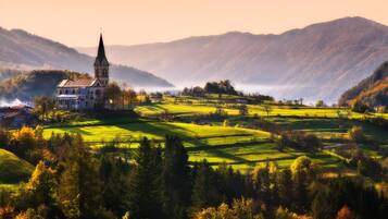 Eine Kirche mit einem hohen Kirchturm, umgeben von sanften grünen Hügeln und einem verschwommenen Bergpanorama.