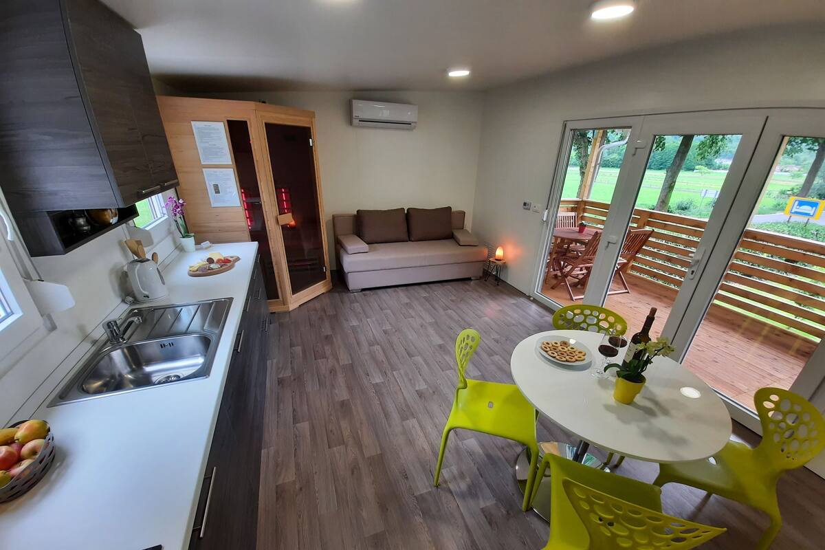 A modern kitchen with a sink, a dining area with a table and chairs, a sofa, and a view of a garden through large glass doors.