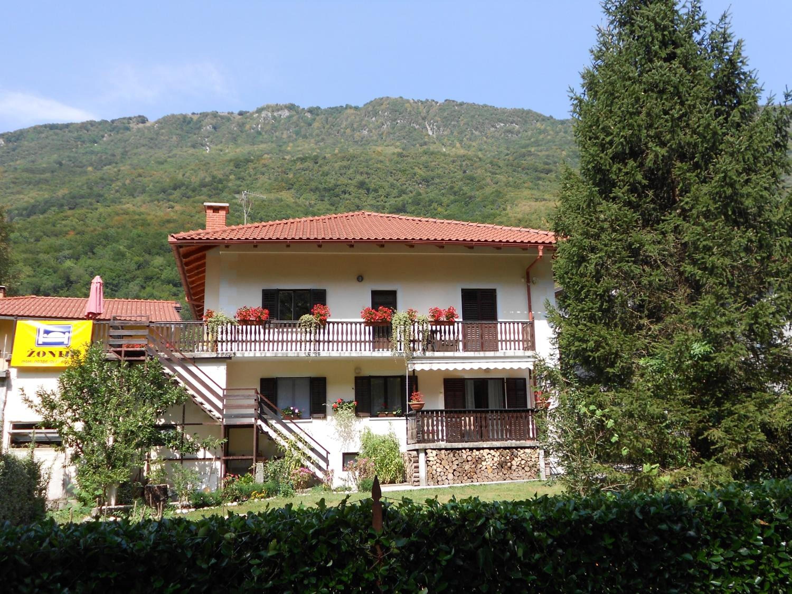 Un edificio de dos pisos con techo de tejas rojas, balcones con jardineras y un paisaje montañoso de fondo.