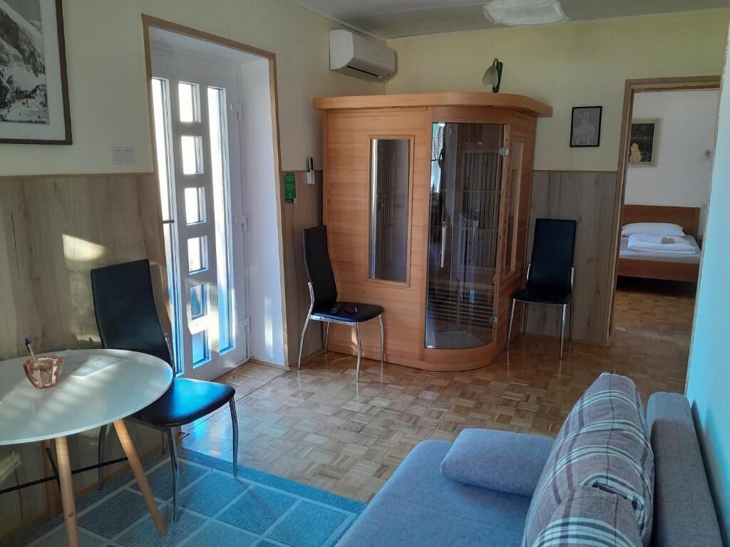 Una stanza con un tavolo rotondo, due sedie, un divano, un piccolo mobiletto in legno e un letto con copripiumino bianco.