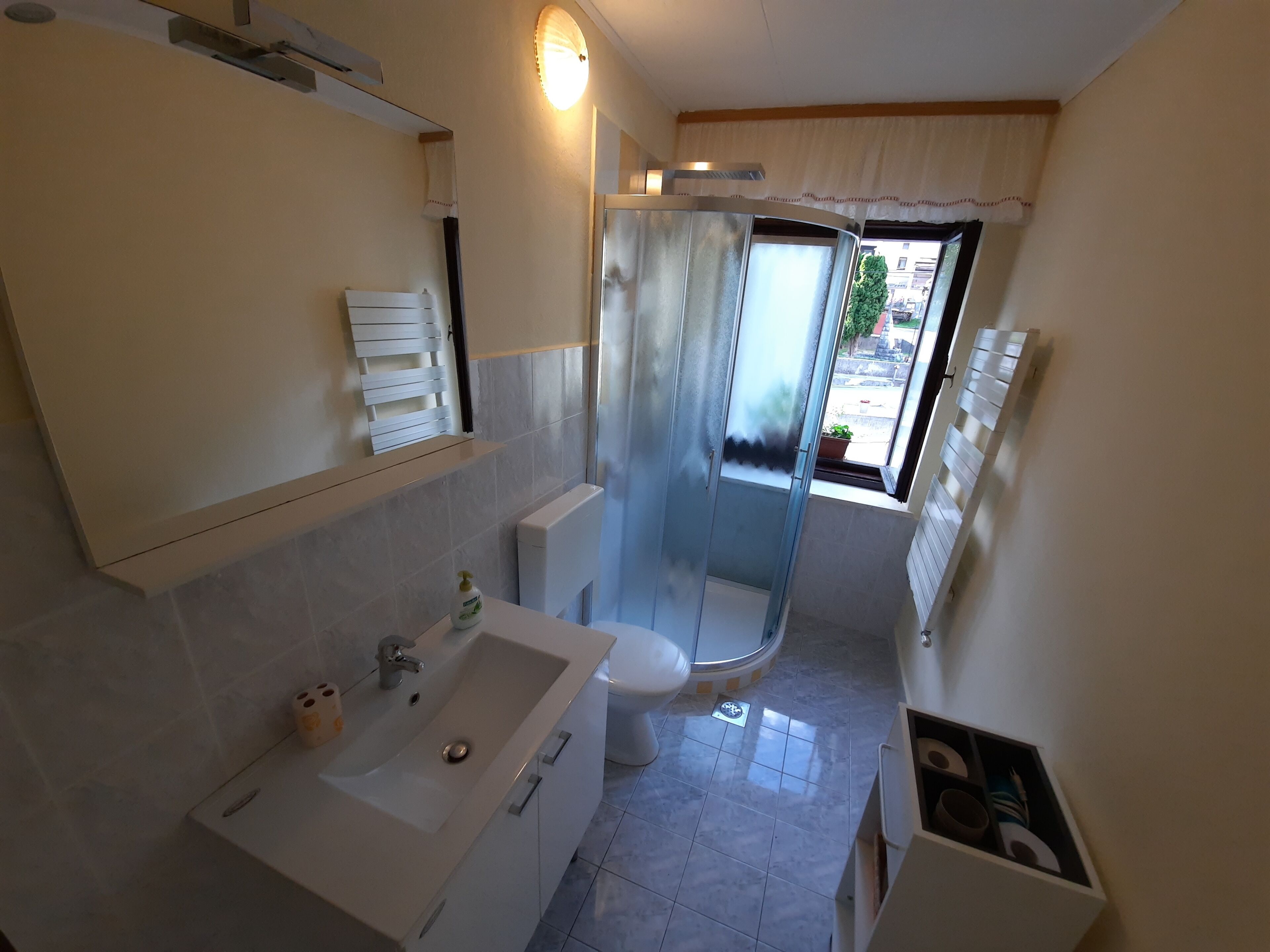 A bathroom with a white sink, toilet, and shower. There is a window with a view of the outside.