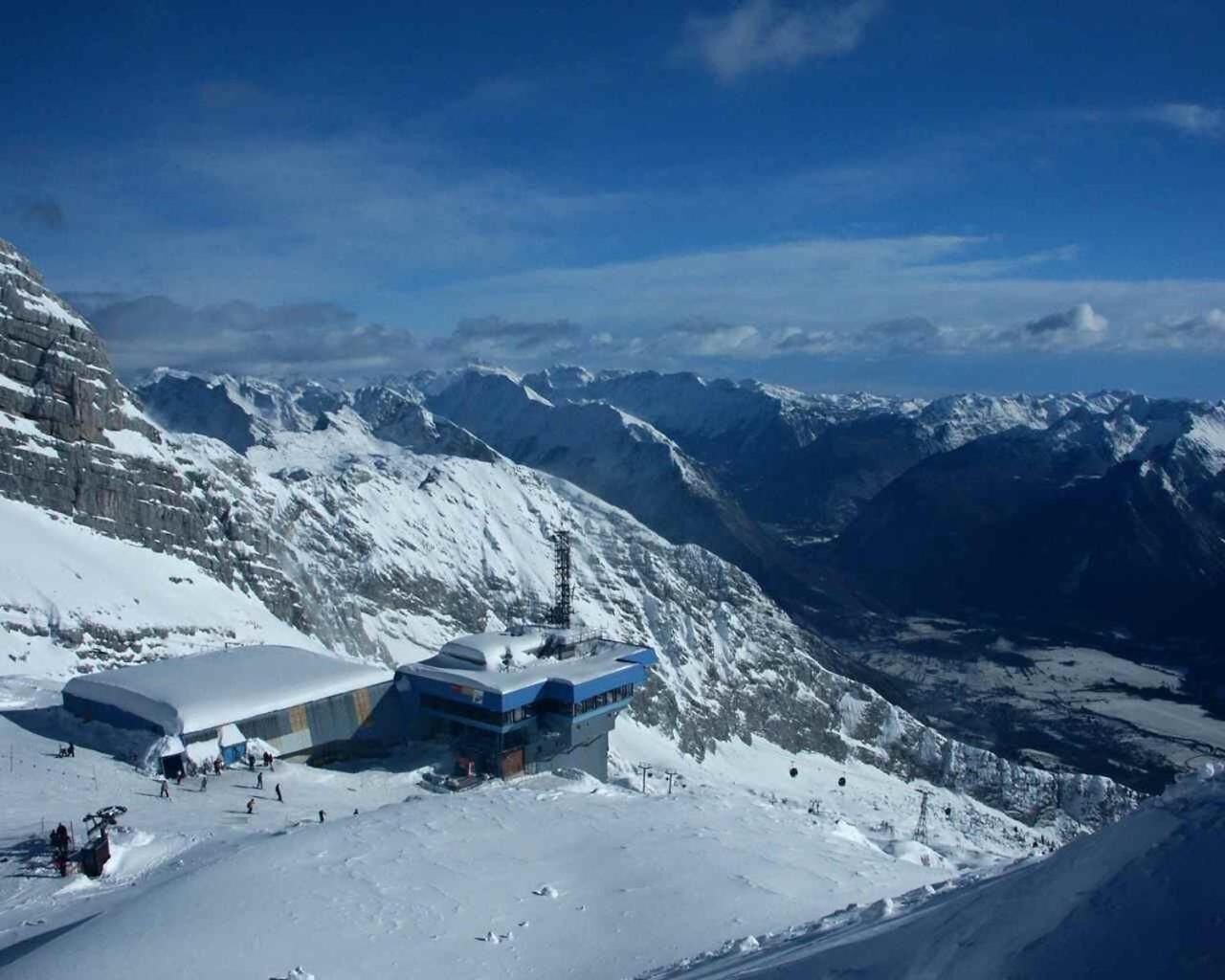 Montanha coberta de neve com uma estação de esqui, destacando um edifício com fachada de vidro e um teleférico em primeiro plano.
