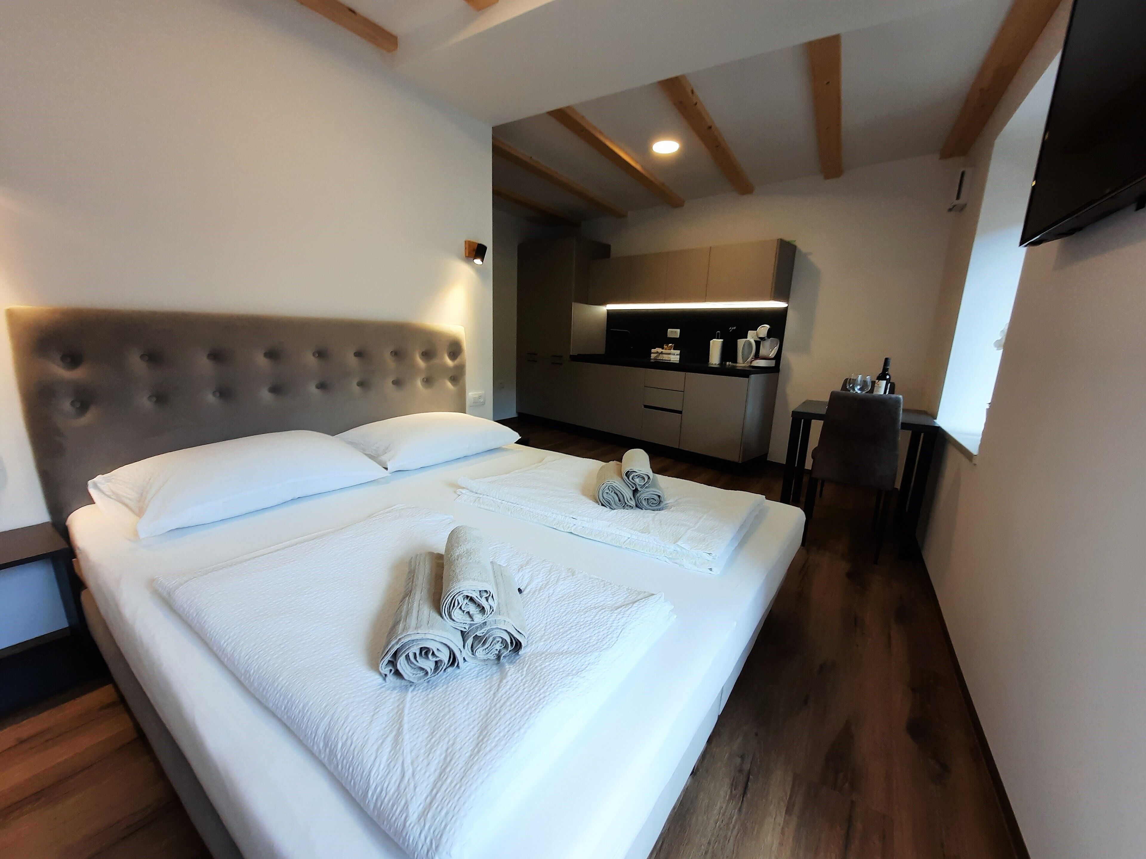Habitación de hotel con una cama grande, cabecera, una pequeña kitchenette, televisor y vigas de madera en el techo.