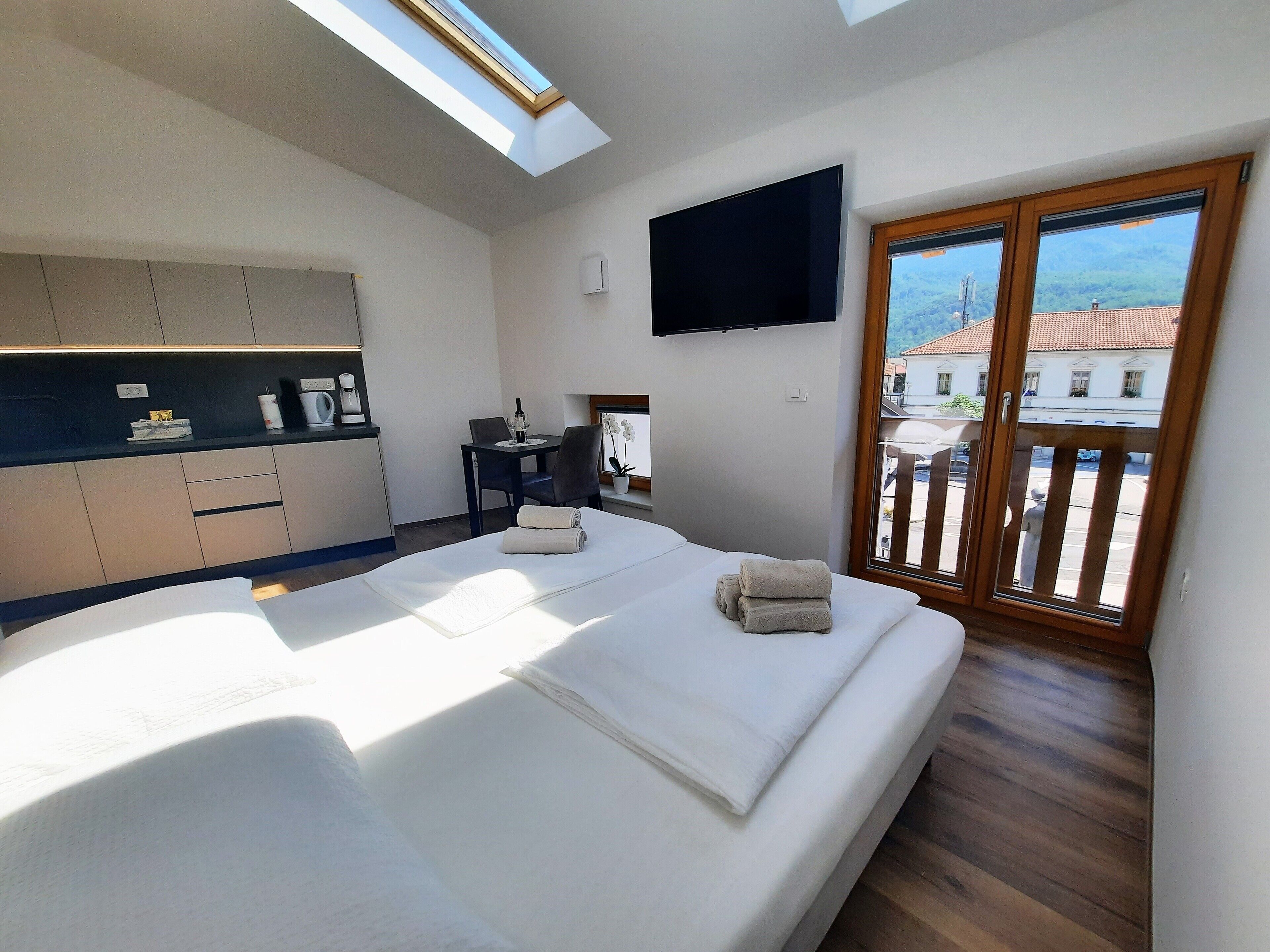 A modern hotel room with a bed, a desk, a TV, and a balcony with a view of buildings.
