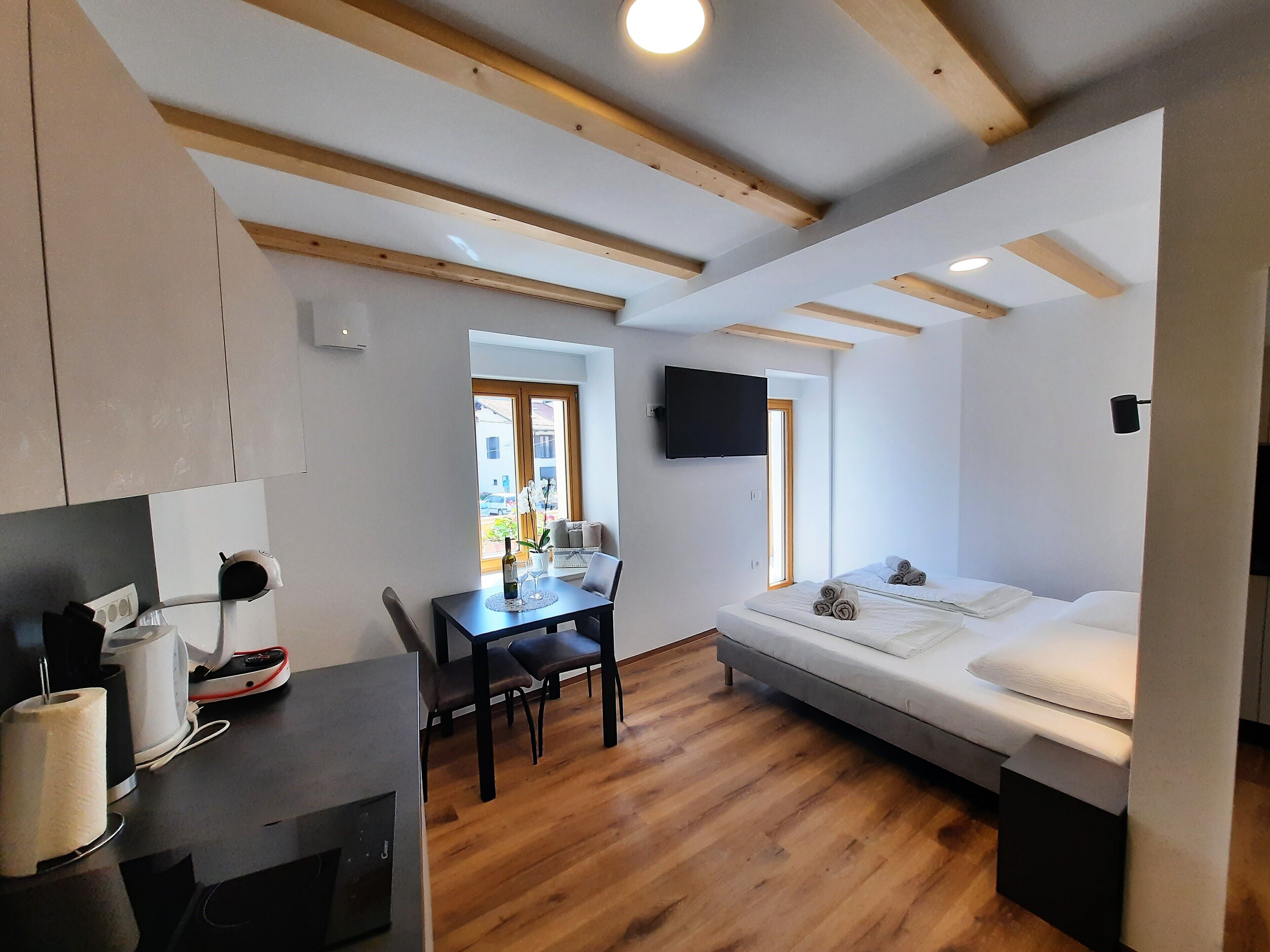 A compact room with a bed, a dining table with chairs, a kitchen area, and a TV mounted on the wall.