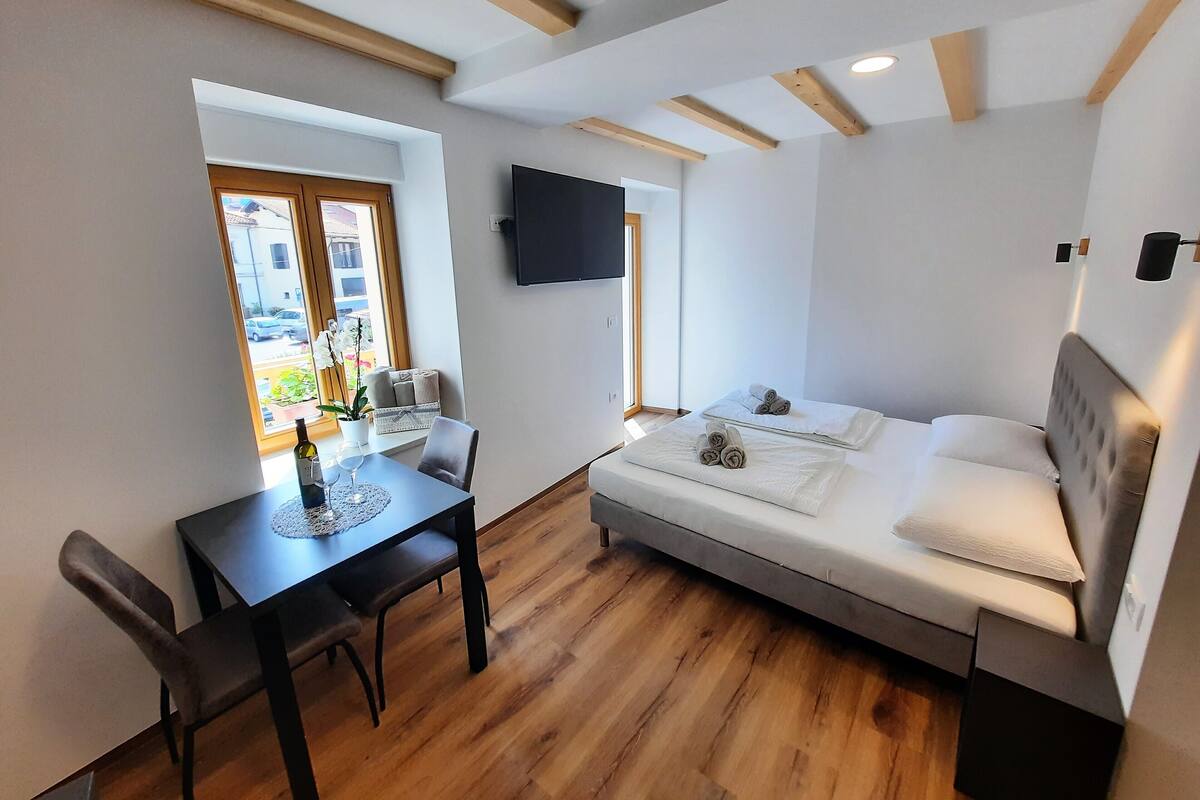 A compact room with a bed, a dining table with chairs, a TV mounted on the wall, and a window with a view of the outside.