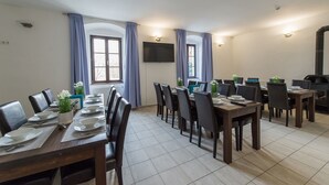 A dining area with multiple tables set for meals, large windows with curtains, a flat-screen TV mounted on the wall, and a wood-burning stove.