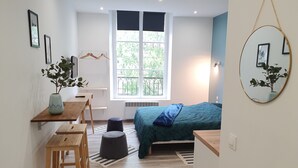 Una habitación compacta con cama, escritorio, silla, un taburete pequeño, una planta en maceta y ventana con vista a follaje.