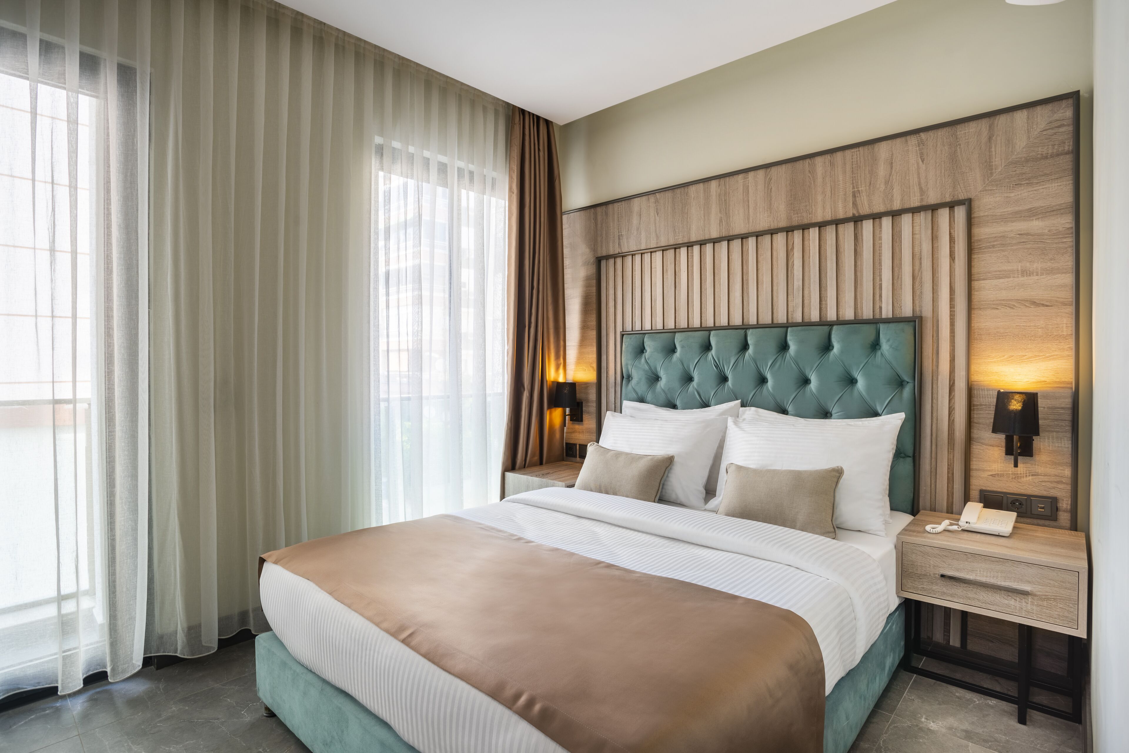 A hotel room with a large bed, a wooden headboard, a nightstand with a lamp, and sheer curtains.