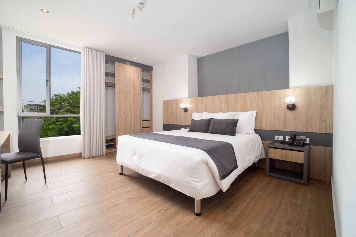 A modern hotel room with a large bed, a desk, and a chair. There is a window with a view of greenery outside.