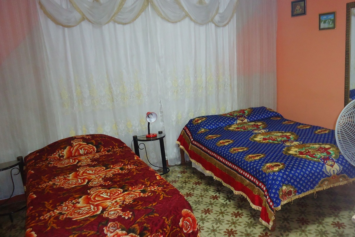 A room with two beds, one with a red floral bedspread and the other with a blue patterned bedspread. There is a standing fan and a small table with a mirror and a lamp.
