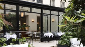 Een terras met tafels en stoelen, omgeven door plantenbakken en een grote raam waardoor het straatbeeld zichtbaar is.