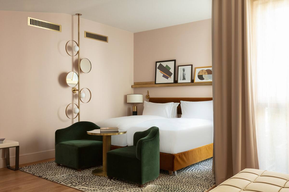Hotelkamer met een groot bed, twee groene fauteuils, een klein rond tafeltje, een bedlampje en kunst aan de muur.