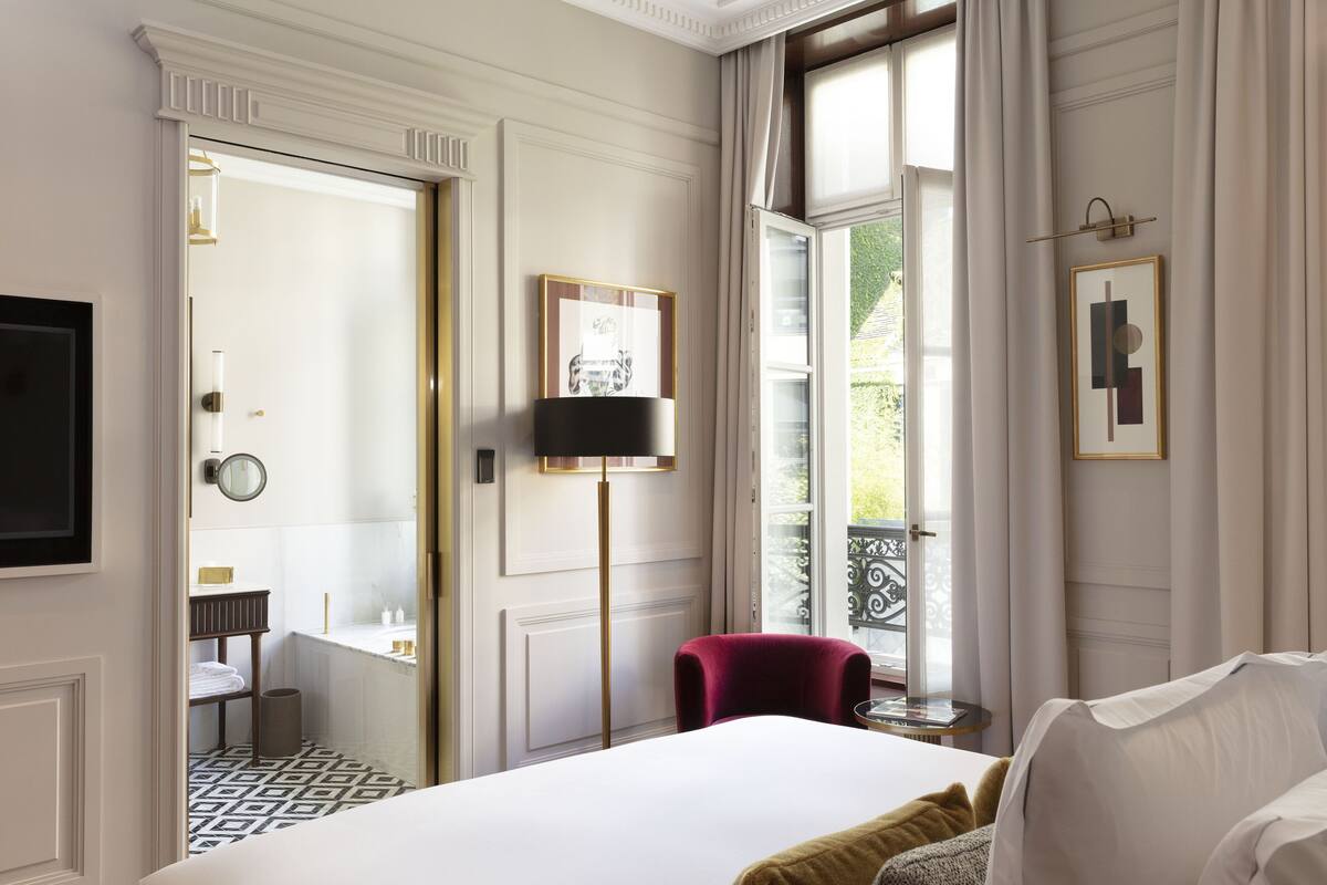 Hotelkamer met een groot bed, een rode fauteuil, een vloerlamp en uitzicht op het buitenlands landschap door een raam.