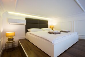 A neatly made bed with white linens and two folded towels, a bedside lamp, and an air conditioning unit on the wall.