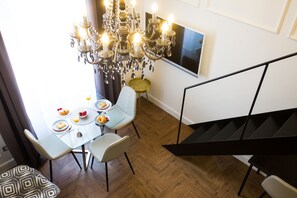 A dining area with a glass table, white chairs, a chandelier, and a TV mounted on the wall.