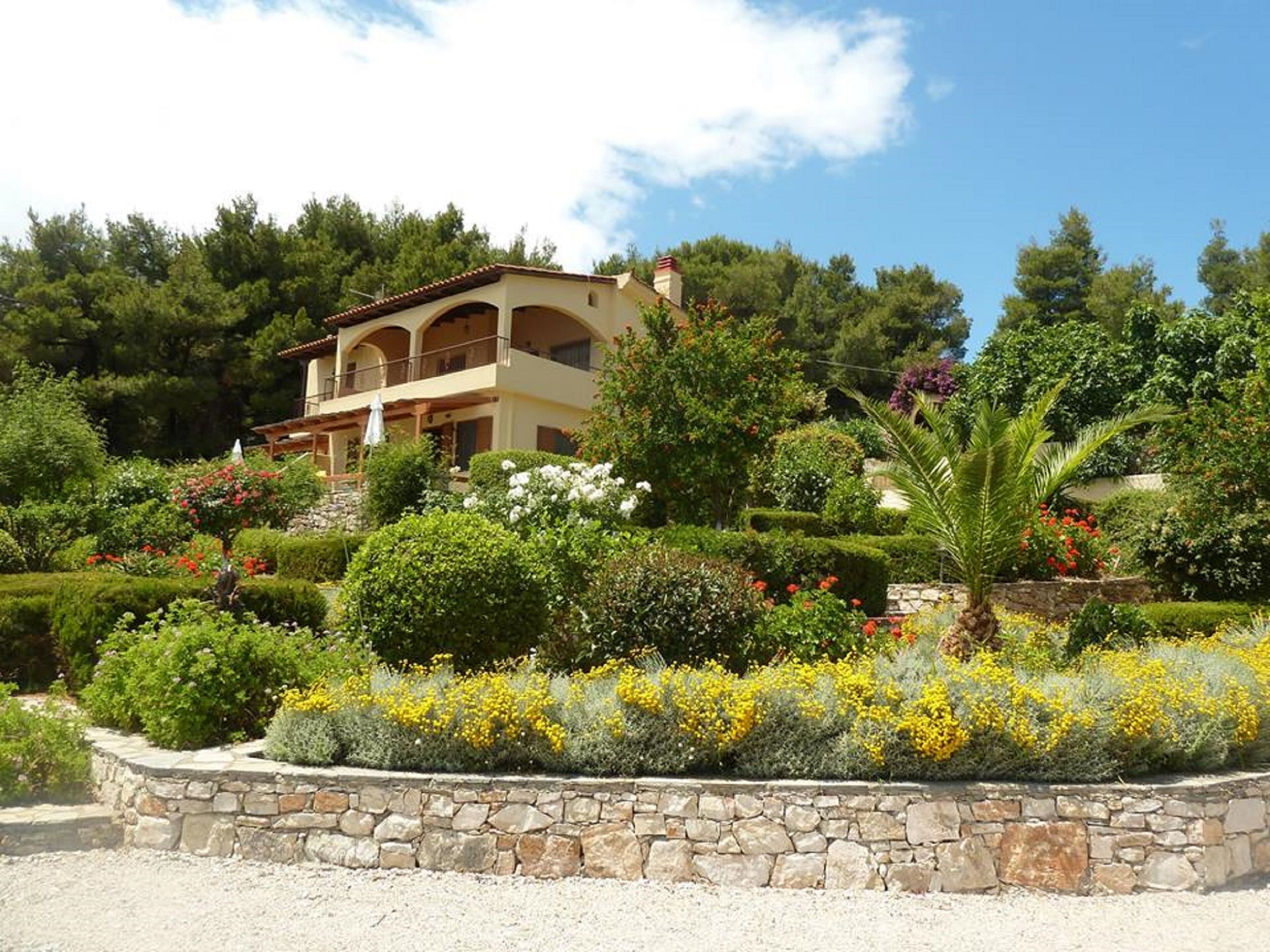 Un immeuble de deux étages, doté d’une façade en pierre et de nombreux balcons, entouré de jardins aménagés avec divers arbustes et plantes à fleurs.