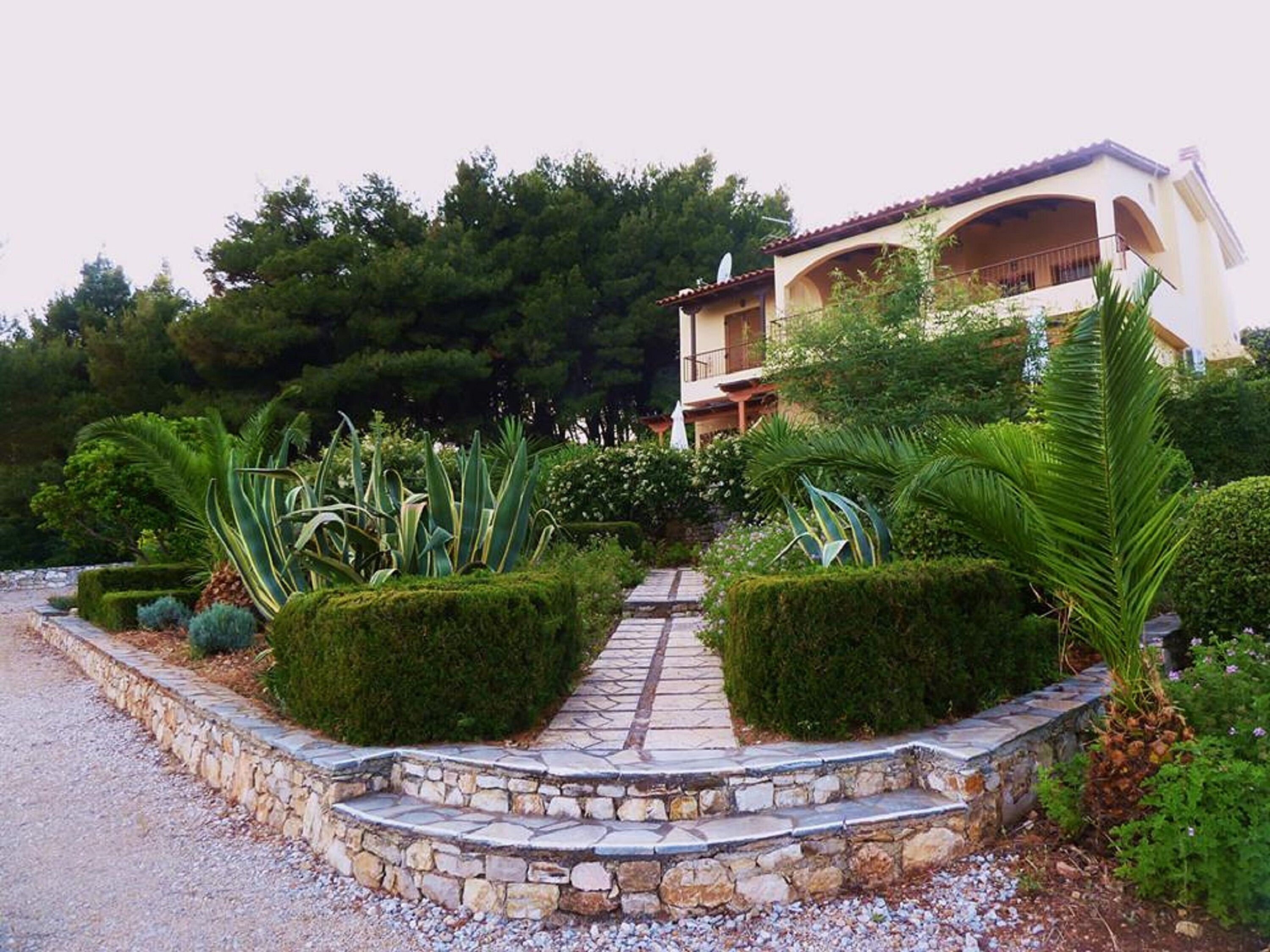 A building with a stone pathway leading to a gated entrance, surrounded by well-maintained hedges and tropical plants.