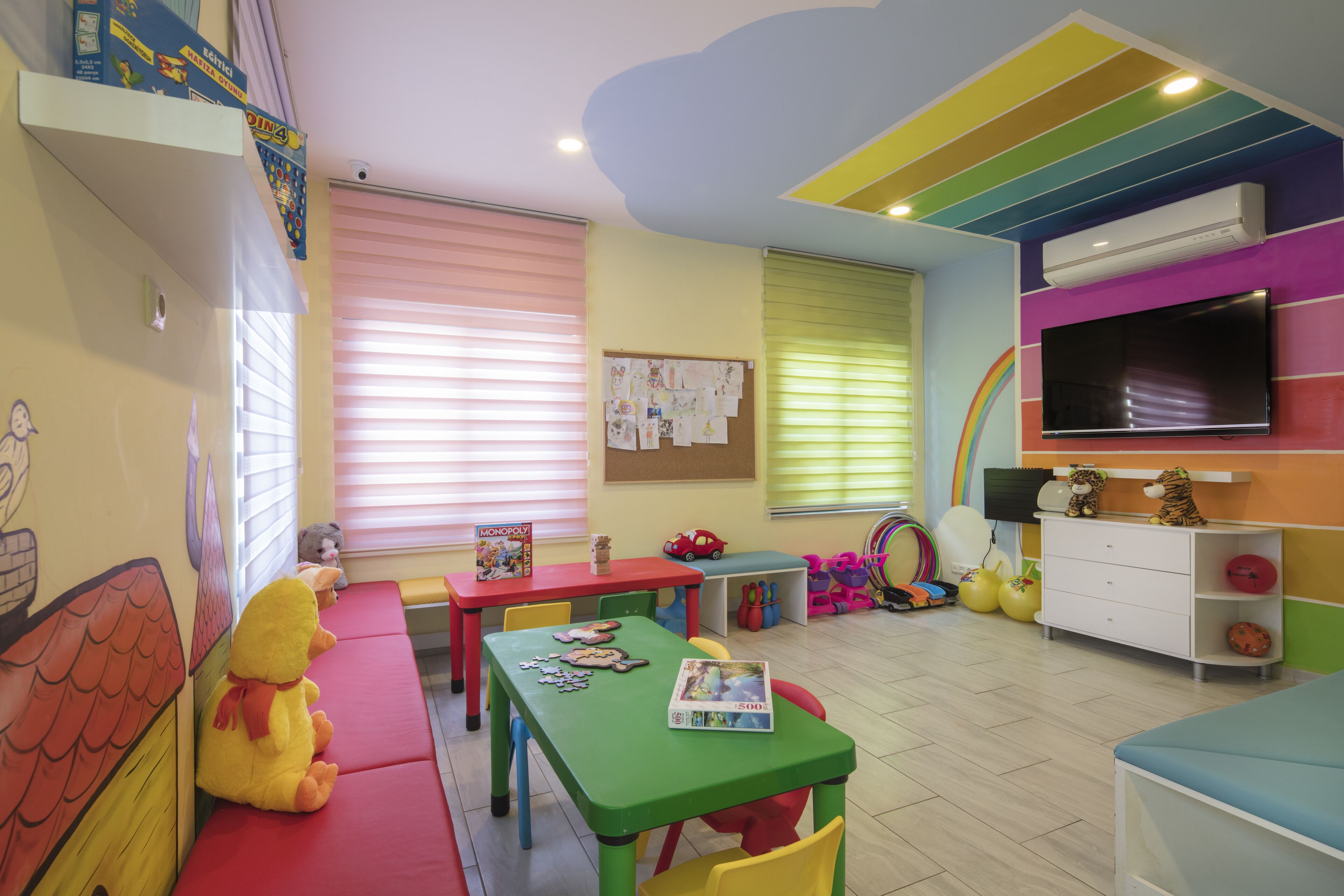 A brightly colored children's playroom with a green table, red and yellow chairs, and a television on the wall.