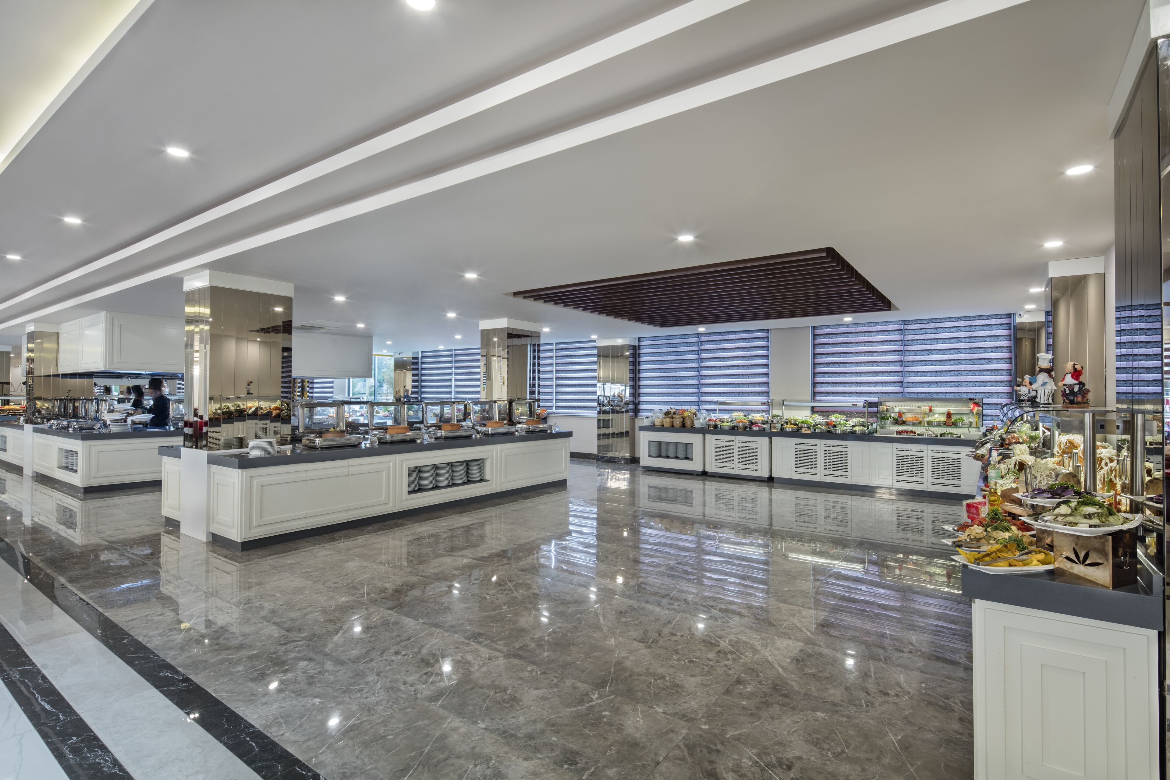 A spacious, modern hotel lobby with a polished marble floor, a central island with food displays, and a large window with blinds.