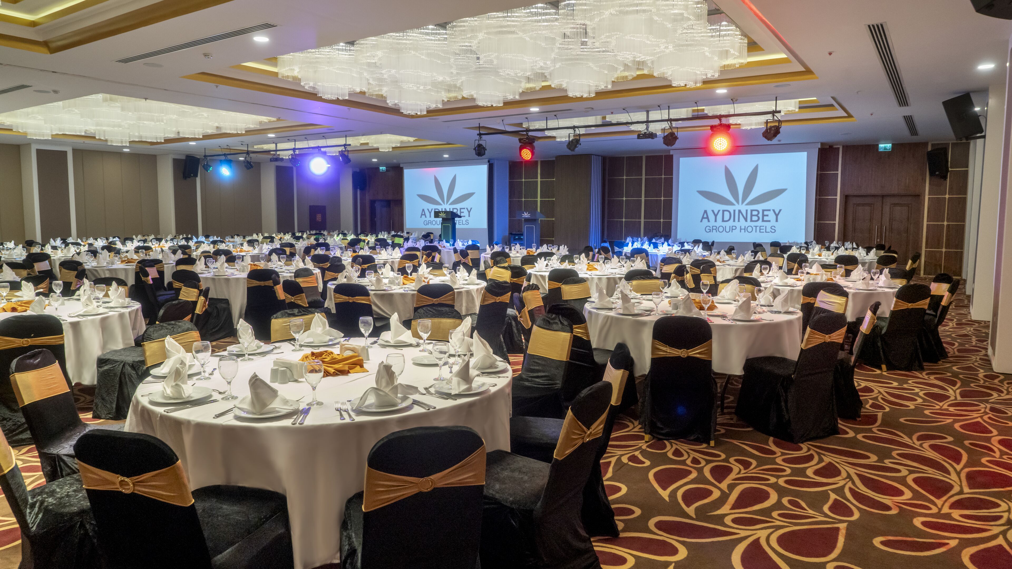 A large conference room with round tables, black chairs with gold sashes, and a chandelier.