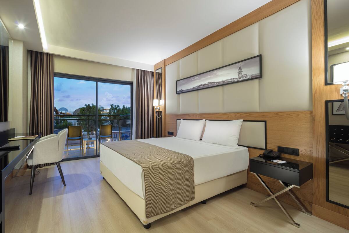A hotel room with a bed, a desk, a chair, a TV, a bedside table, and a view of the cityscape.
