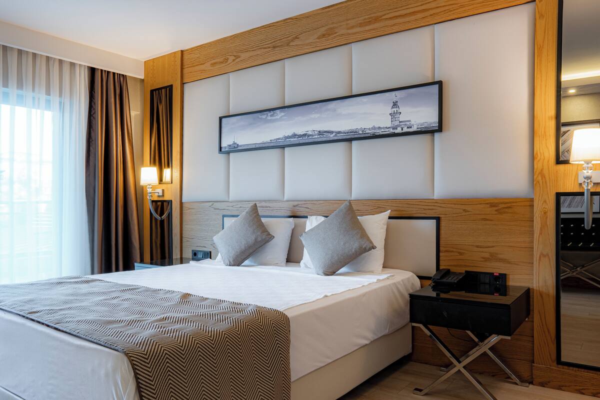 A hotel room with a large bed, a headboard with a cityscape picture, a bedside table with a phone, and a window with curtains.
