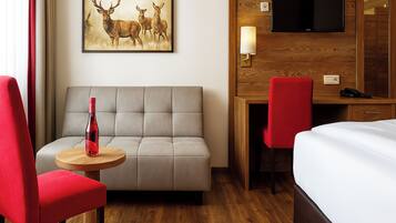 Ein Hotelzimmer mit einer Couch, einem roten Sessel, einem kleinen Tisch mit einer Flasche, einem Schreibtisch mit Stuhl, einem an einer Holzvertäfelung montierten Fernseher und einem Bett.