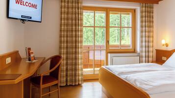 Habitación de hotel con cama, escritorio, silla, televisor mostrando “BIENVENIDOS” y ventana con cortinas.