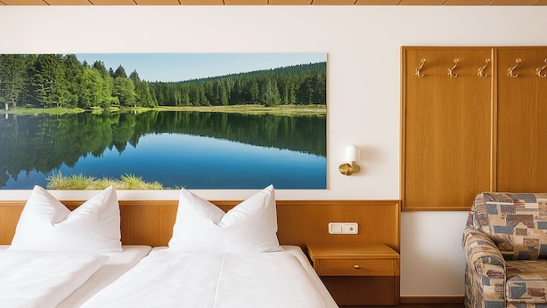 A bedroom with a large bed, a nightstand, a wooden wardrobe, a patterned armchair, and a wall-mounted painting of a lake. - Hotel zur Linde (Lohberg)